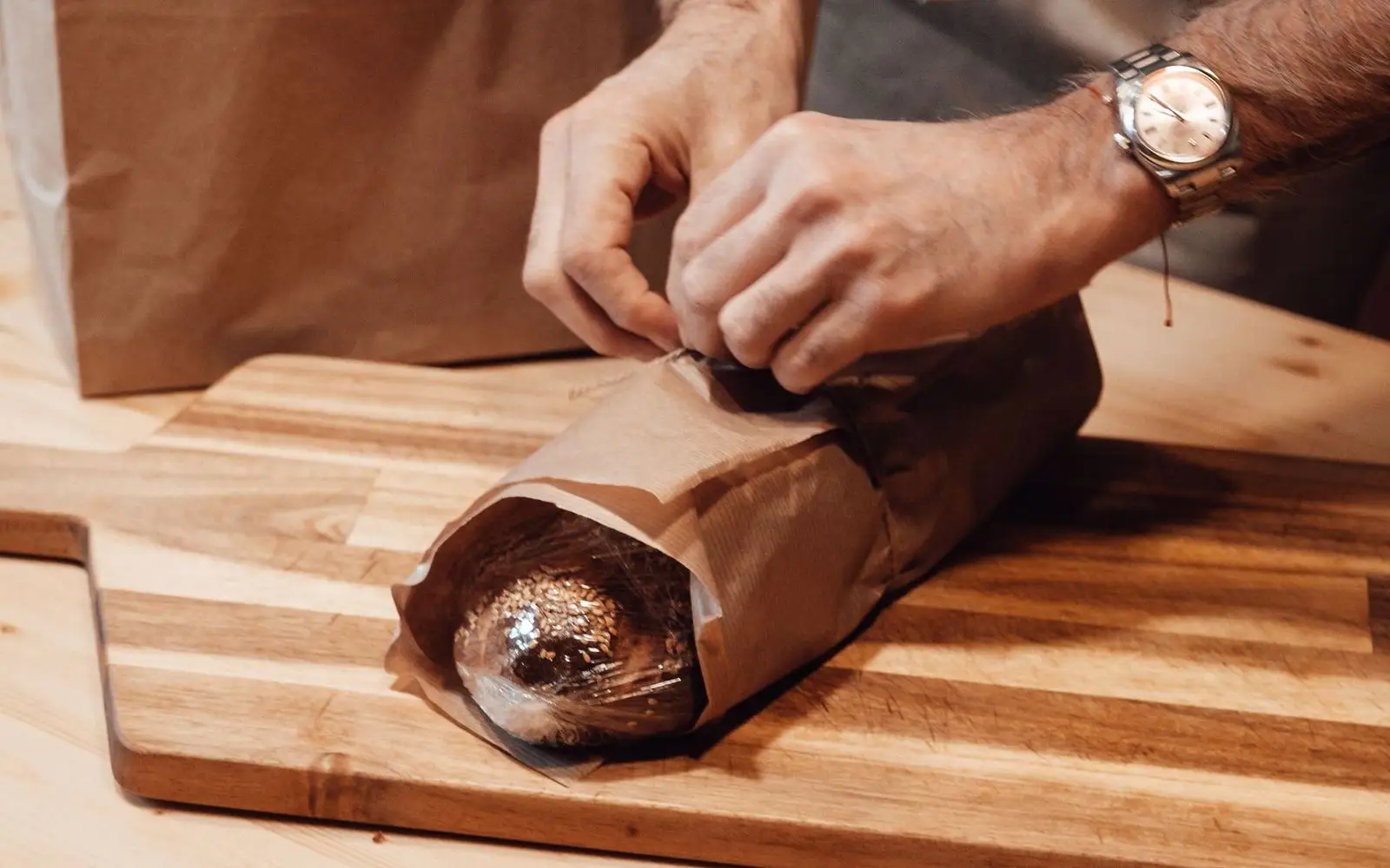Hands wrapping a loaf of bread in brown paper on a wooden board